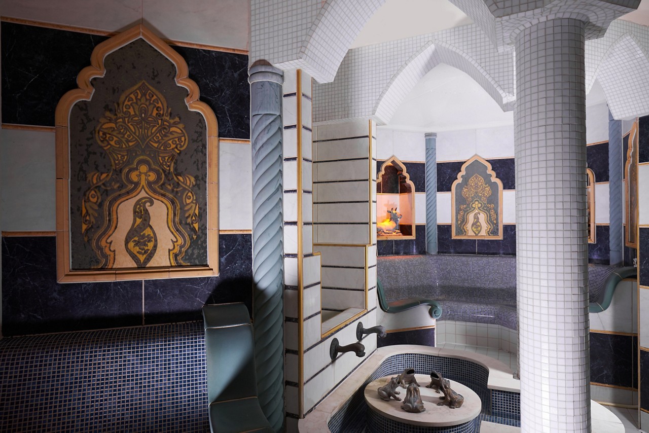 Ornate tiled hammam room—curved bench seating lines the walls, water taps feed a small central basin with figurines—surrounded by arched niches, patterned panels, spiral columns, and vaulted, mosaic-covered ceilings.